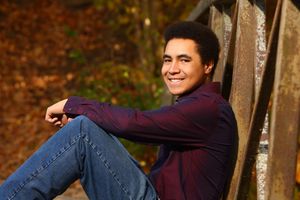 Young man with afro hair smiling, sitting outdoors in autumn foliage wearing a maroon shirt and jeans.