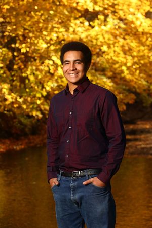 Smiling young man in burgundy shirt and jeans poses outdoors with vibrant golden autumn foliage background.