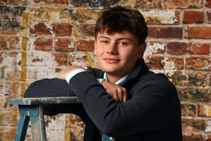 Young man in dark jacket posing confidently against rustic brick wall backdrop in a professional portrait photo.