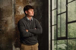 Young man in gray sweater with arms crossed leaning against wall near window.