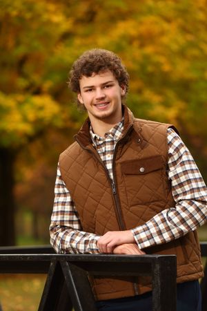 Young man with curly hair smiling in a brown quilted vest and plaid shirt, leaning on a fence with autumn foliage.