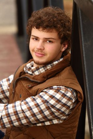 Young man with curly hair wearing a brown vest over plaid shirt, leaning against a dark wall outdoors.