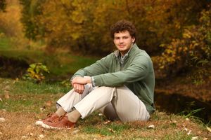 Young man with curly hair sitting outdoors in autumn park wearing green sweater and khaki pants.