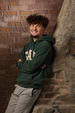 Young man with curly hair smiling, wearing a green CAL hoodie, leaning against a brick wall with arms crossed.