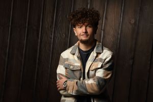 Young man with curly hair in a plaid jacket stands arms crossed against a dark wood wall.