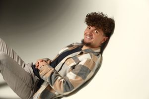 Young man with curly hair smiling, wearing a plaid shirt, relaxing against a light gray background.