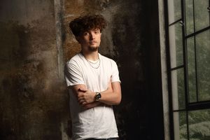 Young man with curly hair in white t-shirt and chain necklace leaning against textured wall with arms crossed.