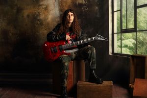 Musician with curly hair playing a red electric guitar, wearing camo pants and leather jacket in a moody studio setting.