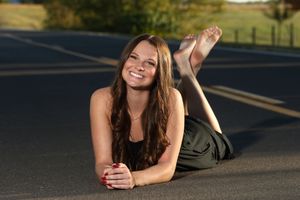 Young woman with long brown hair smiling while lying on a road, wearing a dark dress on a sunny day.