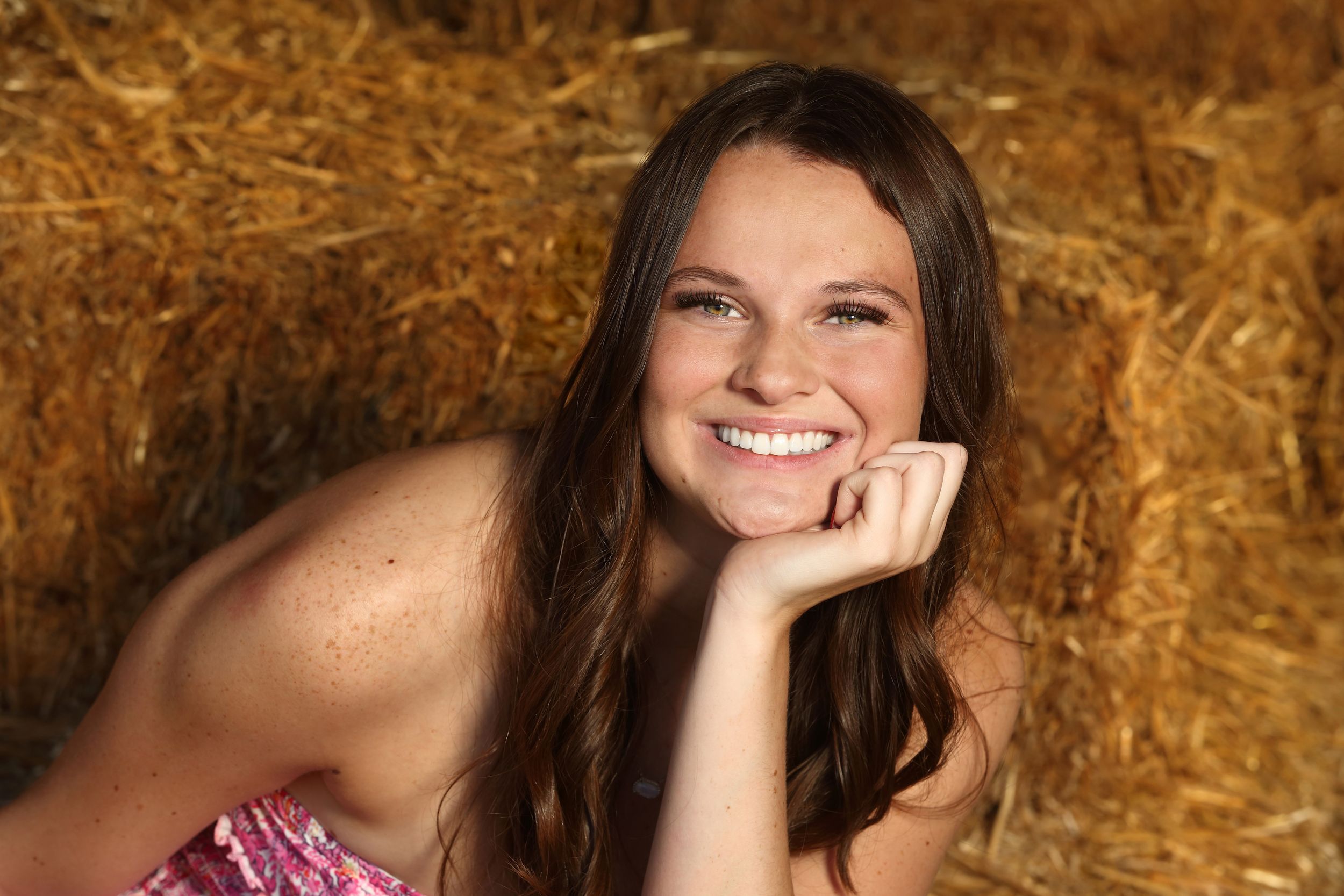 Smiling young woman with long brown hair resting chin on hand, posing in front of golden hay bales.