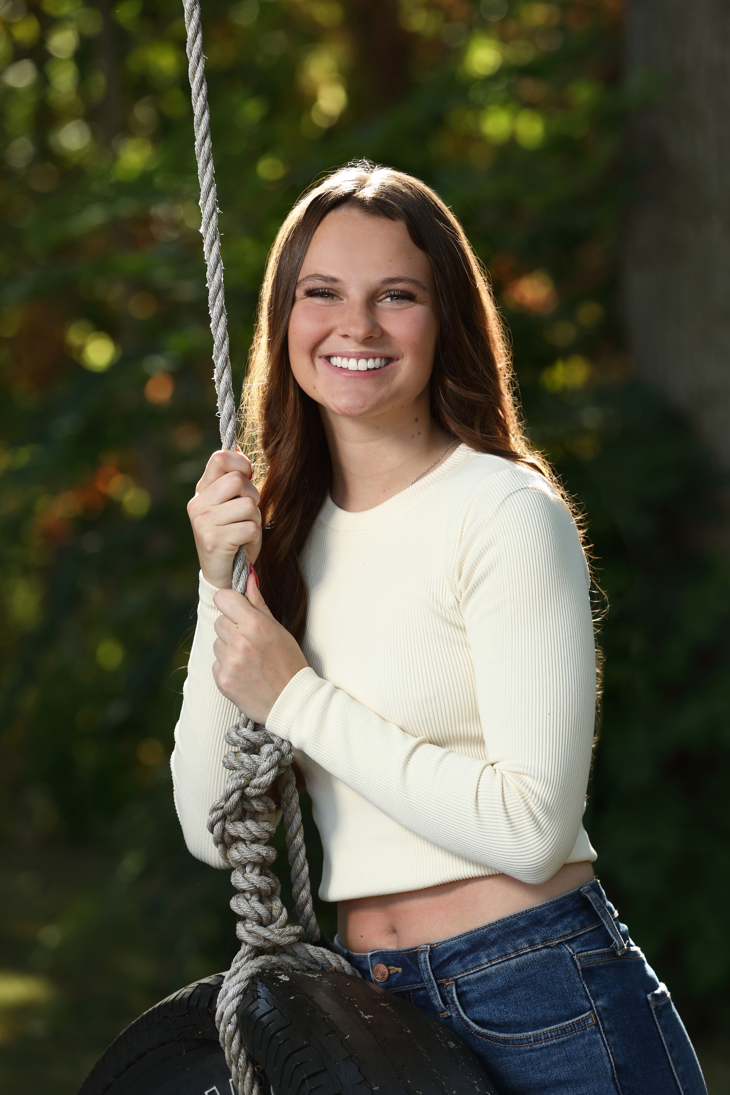 Smiling young woman with long brown hair sitting on a tire swing, wearing a cream crop top and jeans outdoors.