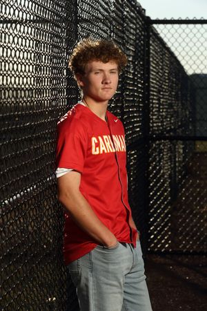 Young male athlete in red Carlman jersey leans against chain-link fence, wearing jeans, in outdoor sports facility.