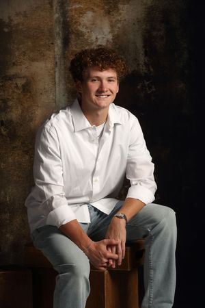 Young man with curly red hair smiling, wearing white shirt and jeans, seated in a studio portrait setting.