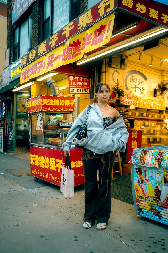 Young woman in gray hoodie holds shopping bag outside vibrant Chinatown storefront with Chinese signs and chestnut vendor.