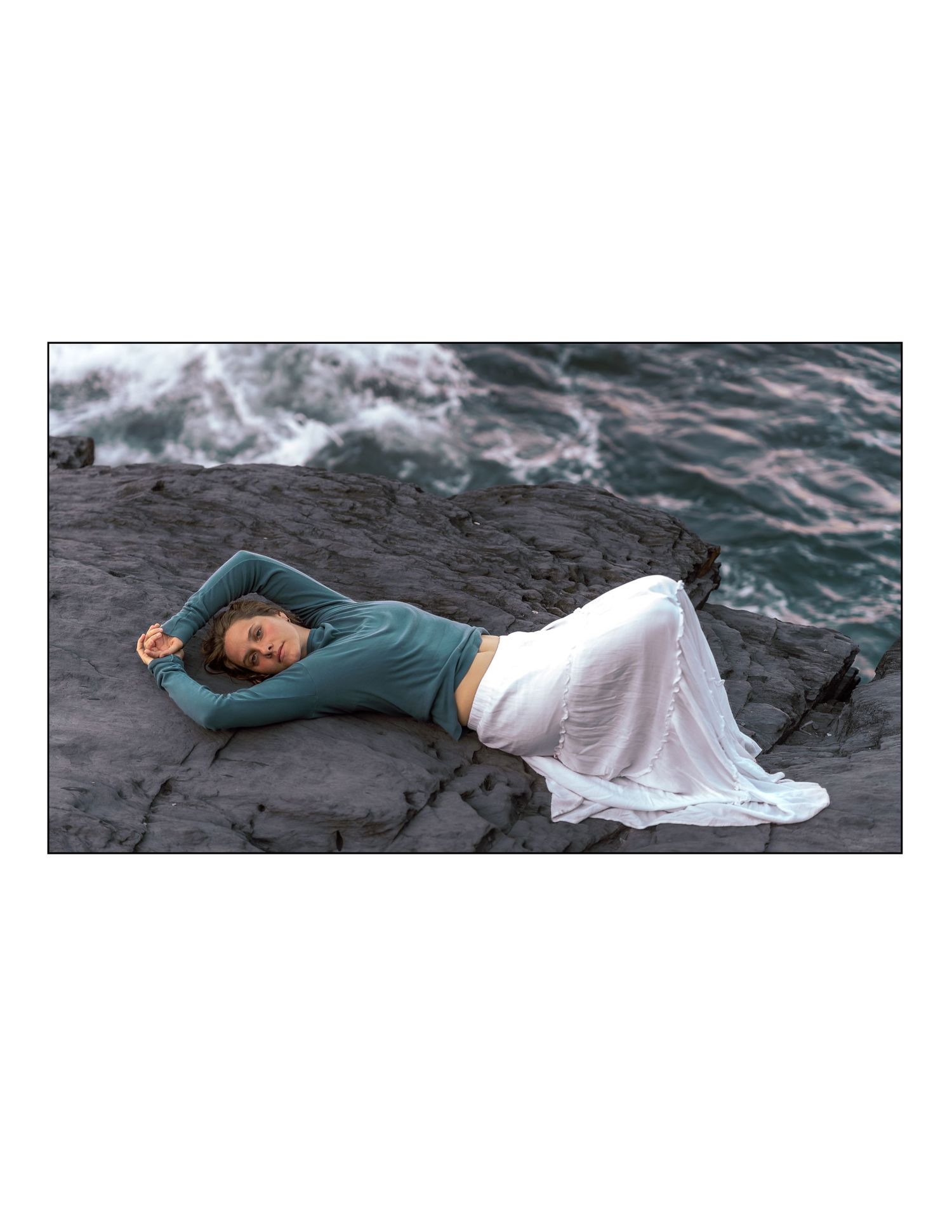 Person in teal top and flowing white skirt lying on dark rocky surface near ocean waves.