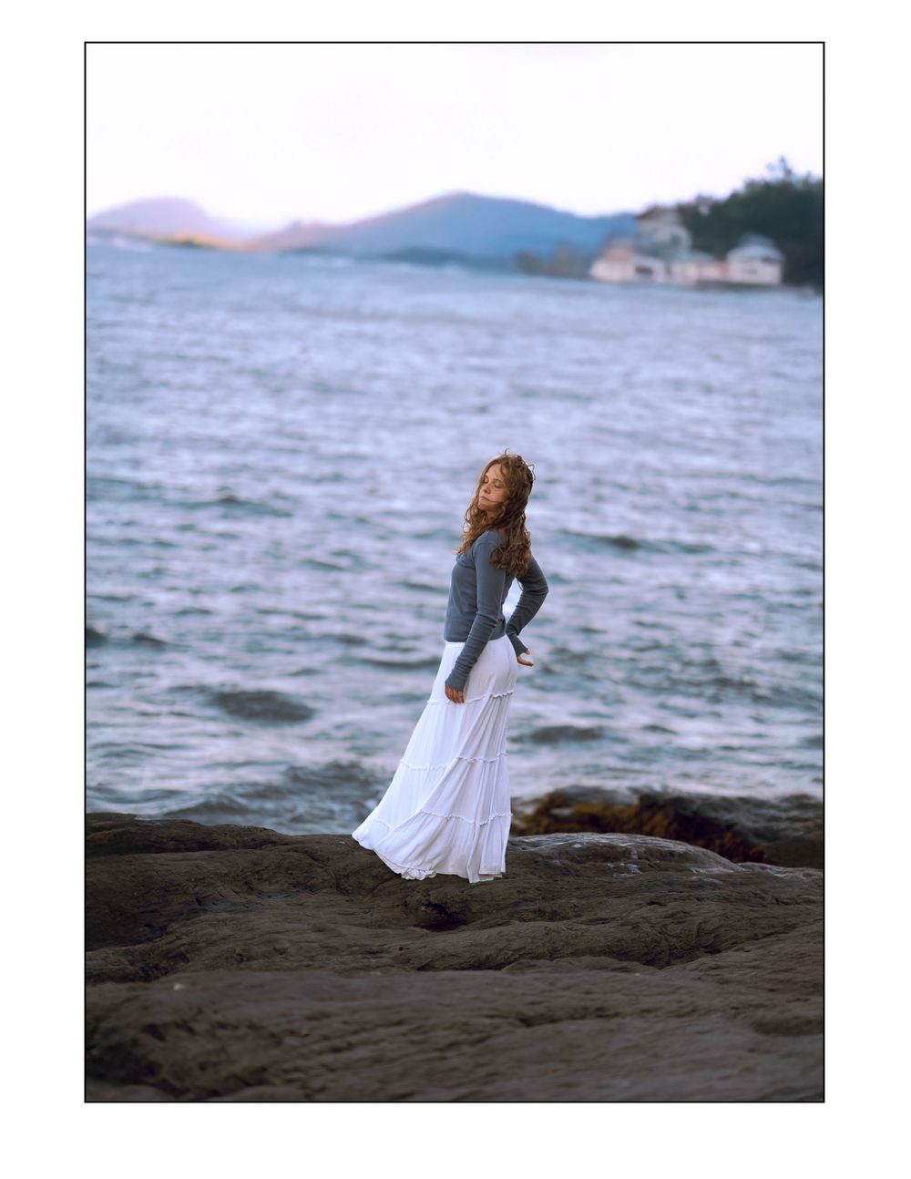 A woman in a flowing white dress stands against a dramatic coastal backdrop during golden hour by the ocean.
