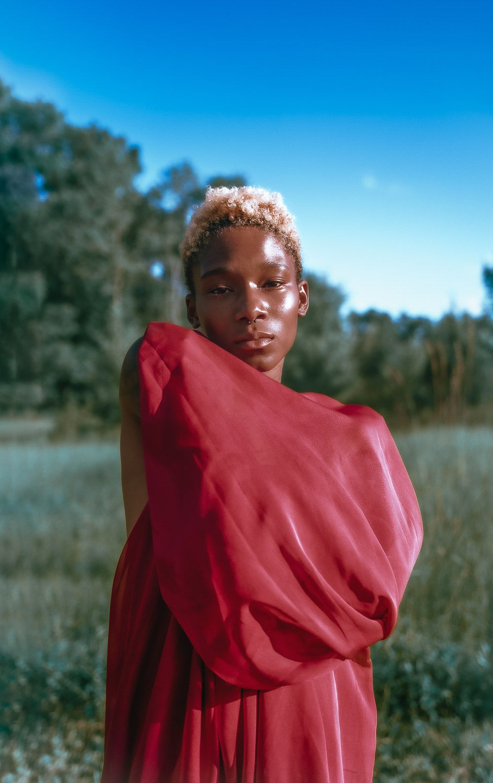 A figure in a flowing red garment stands against a blue sky in a natural outdoor setting.