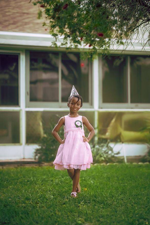 Young Black girl in pink dress and silver party hat stands on green grass in front of a house.