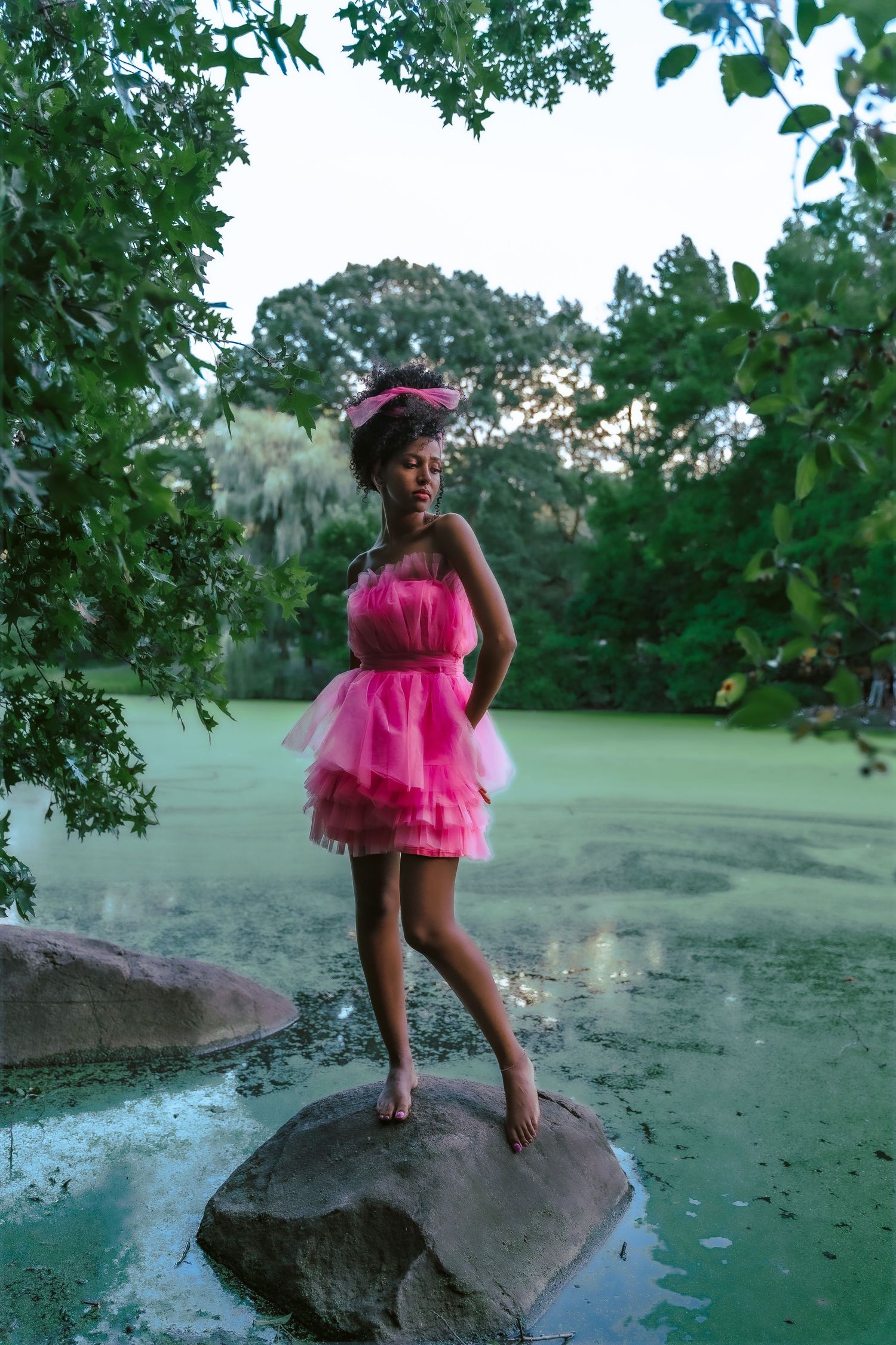 A young woman in a pink tulle dress stands barefoot on a rock by a green pond surrounded by lush trees.