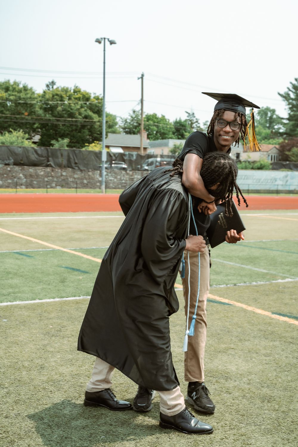 Graduate in cap and gown receives a piggyback ride on a green athletic field during outdoor graduation celebration.