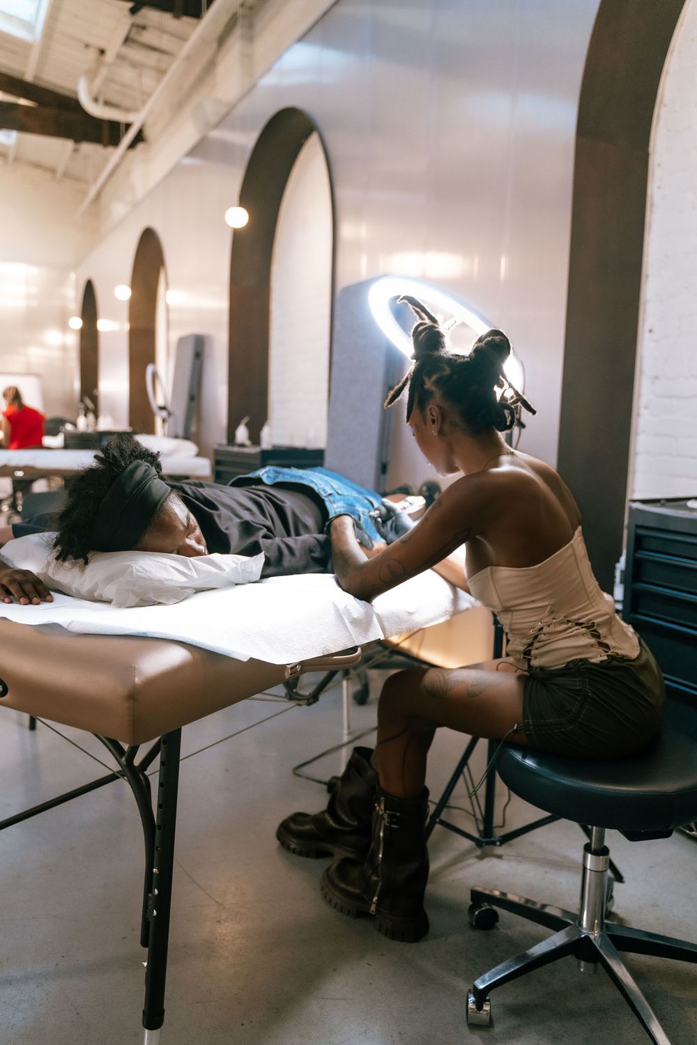Tattoo artist working on a client lying on a table in a modern tattoo studio with arched windows.