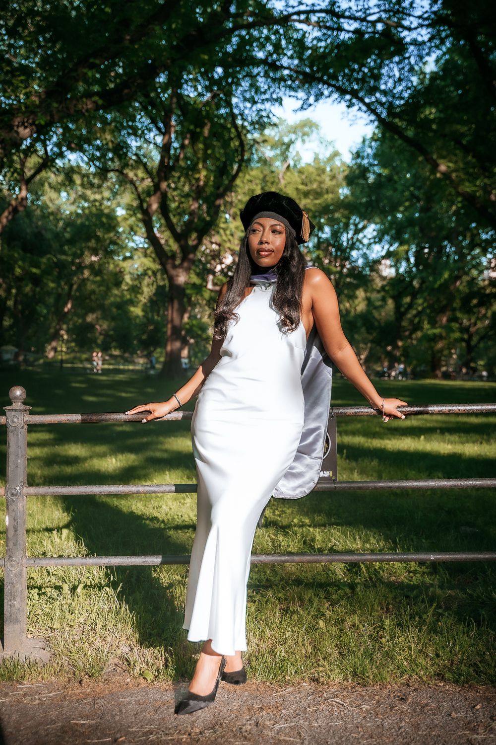 Elegant woman in white dress poses confidently against a rustic fence in a park setting.