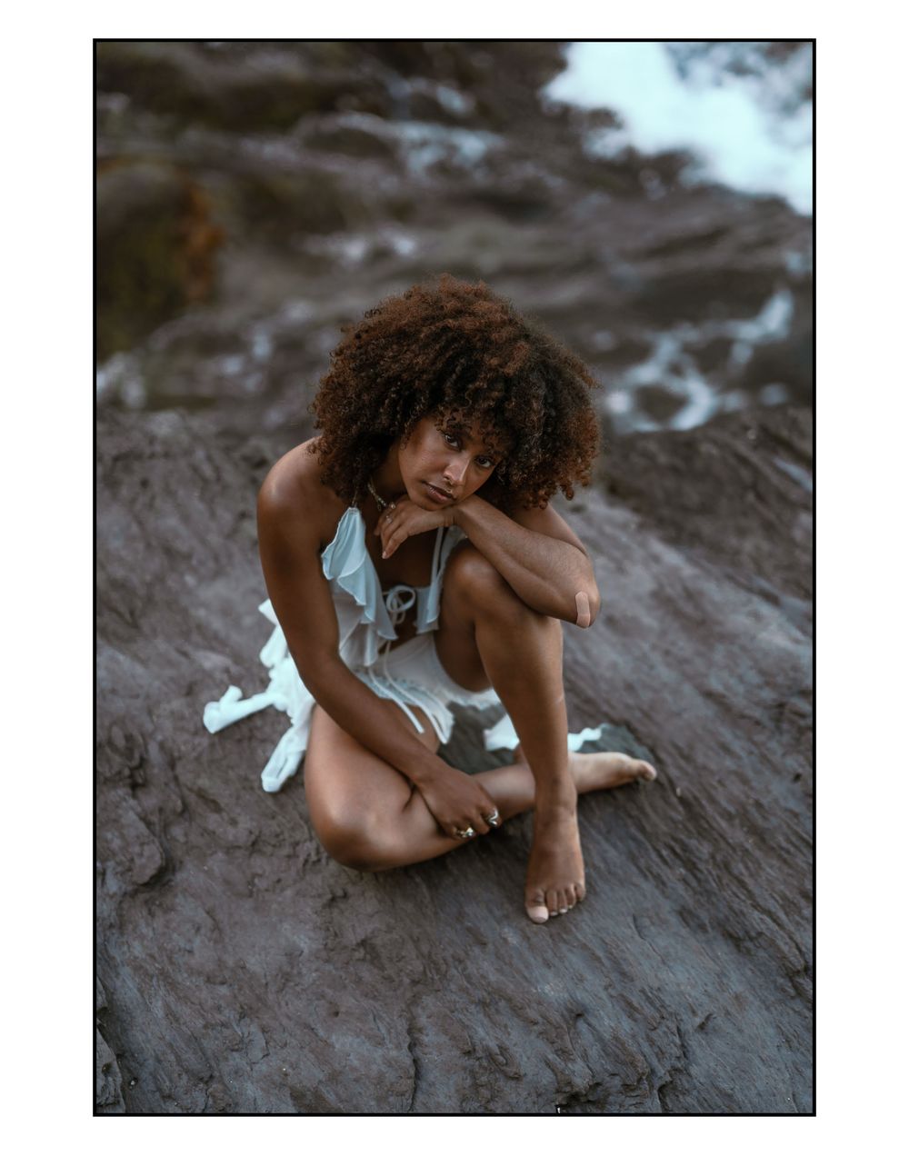 Someone in a flowy white dress sits barefoot on a dark rocky surface during golden hour.