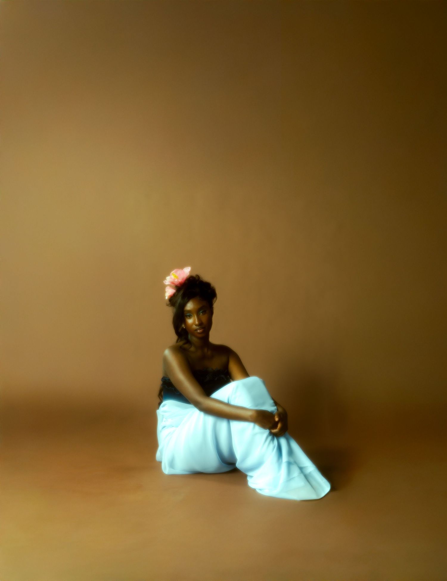 A Black woman in a light blue wrap and pink floral hair accessory poses gracefully on a warm brown studio floor.