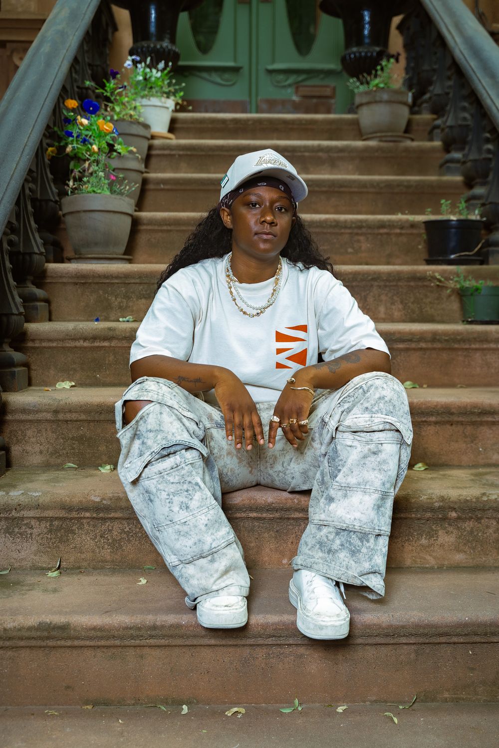 Person sitting on brownstone steps wearing white tee, acid wash cargo pants, white sneakers and cap.