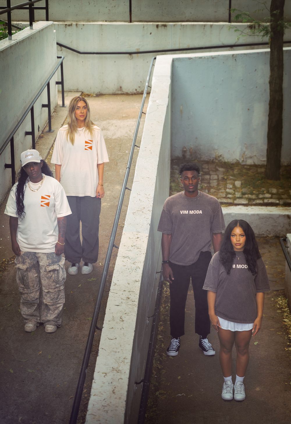 Four young people wearing branded white and grey oversized t-shirts posed on outdoor concrete steps.