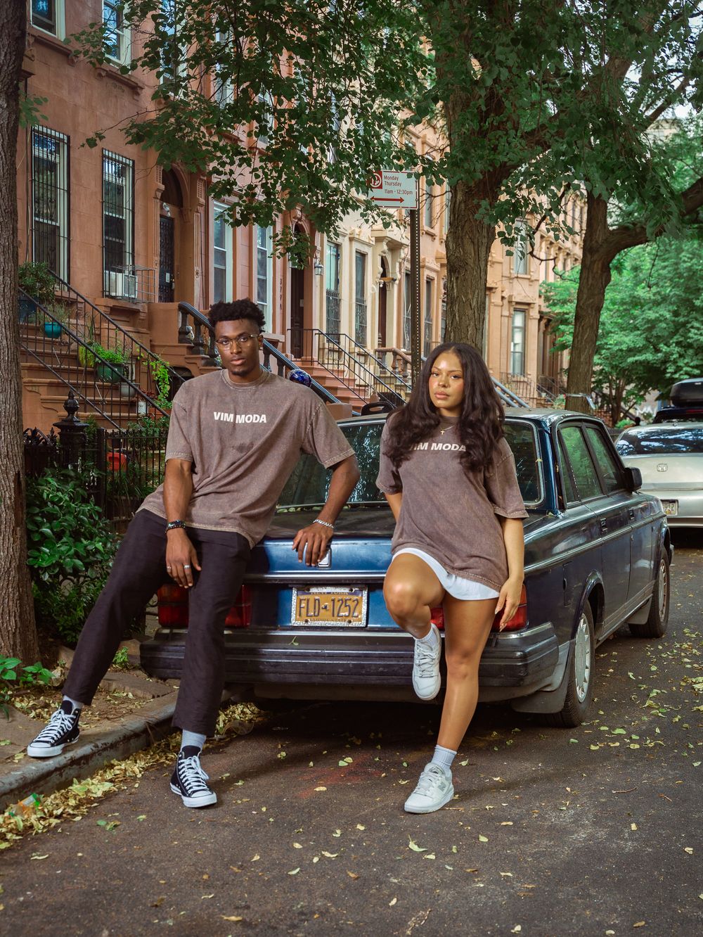 Two models in matching brown VIM MODA tees pose by a vintage car on a Brooklyn street lined with brownstones.