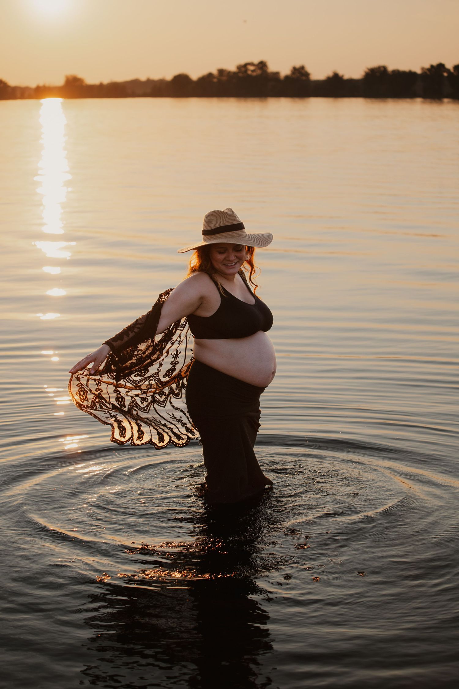 Expectant mother in a hat stands in lake water during golden hour sunset.