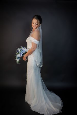 Elegant bridal portrait in white lace gown with flowing train against dark background.
