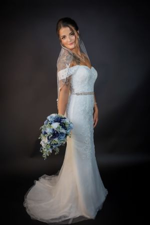 Bride in white lace gown holds blue bouquet during studio photoshoot.