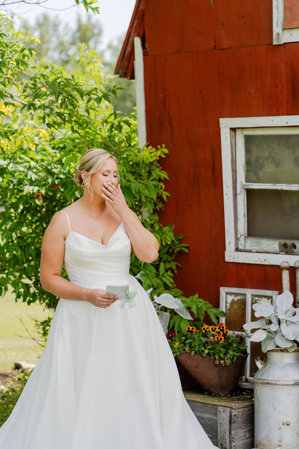 An emotional bride in a white dress stands near a rustic red barn wall during an intimate pre-wedding moment.