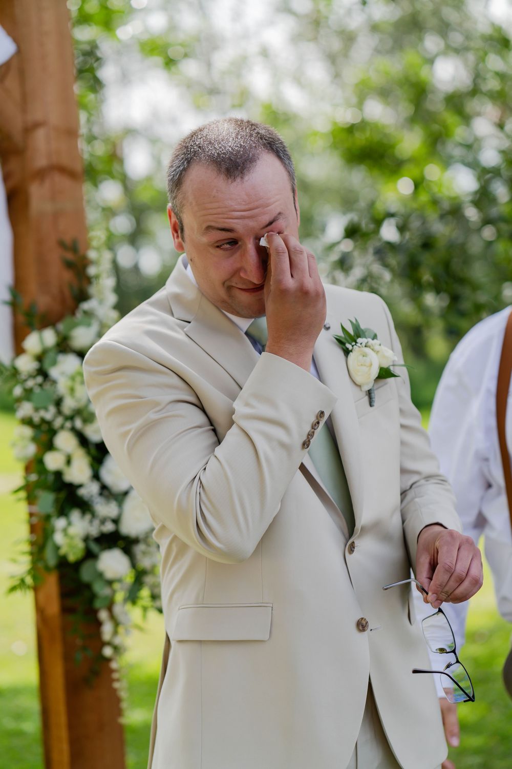 Three photos showing an emotional groom in a cream suit wiping away tears during an outdoor wedding ceremony.