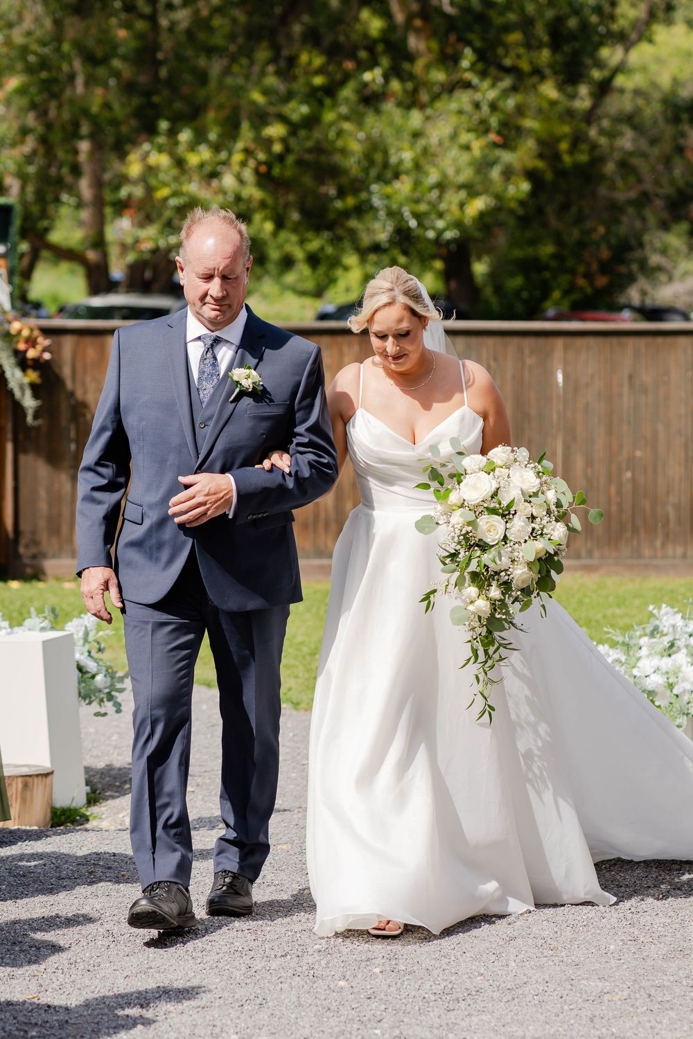 Outdoor wedding ceremony moment showing a person in a navy suit escorting someone in a white wedding gown down a path.