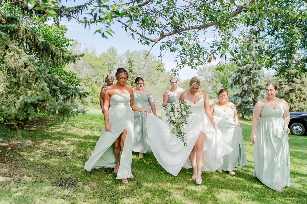 A group in white dresses poses playfully under trees in a sunlit outdoor setting for wedding photos.
