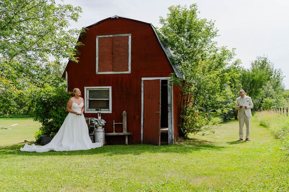 A rustic red barn serves as a backdrop for an outdoor wedding photo session in a lush green field.