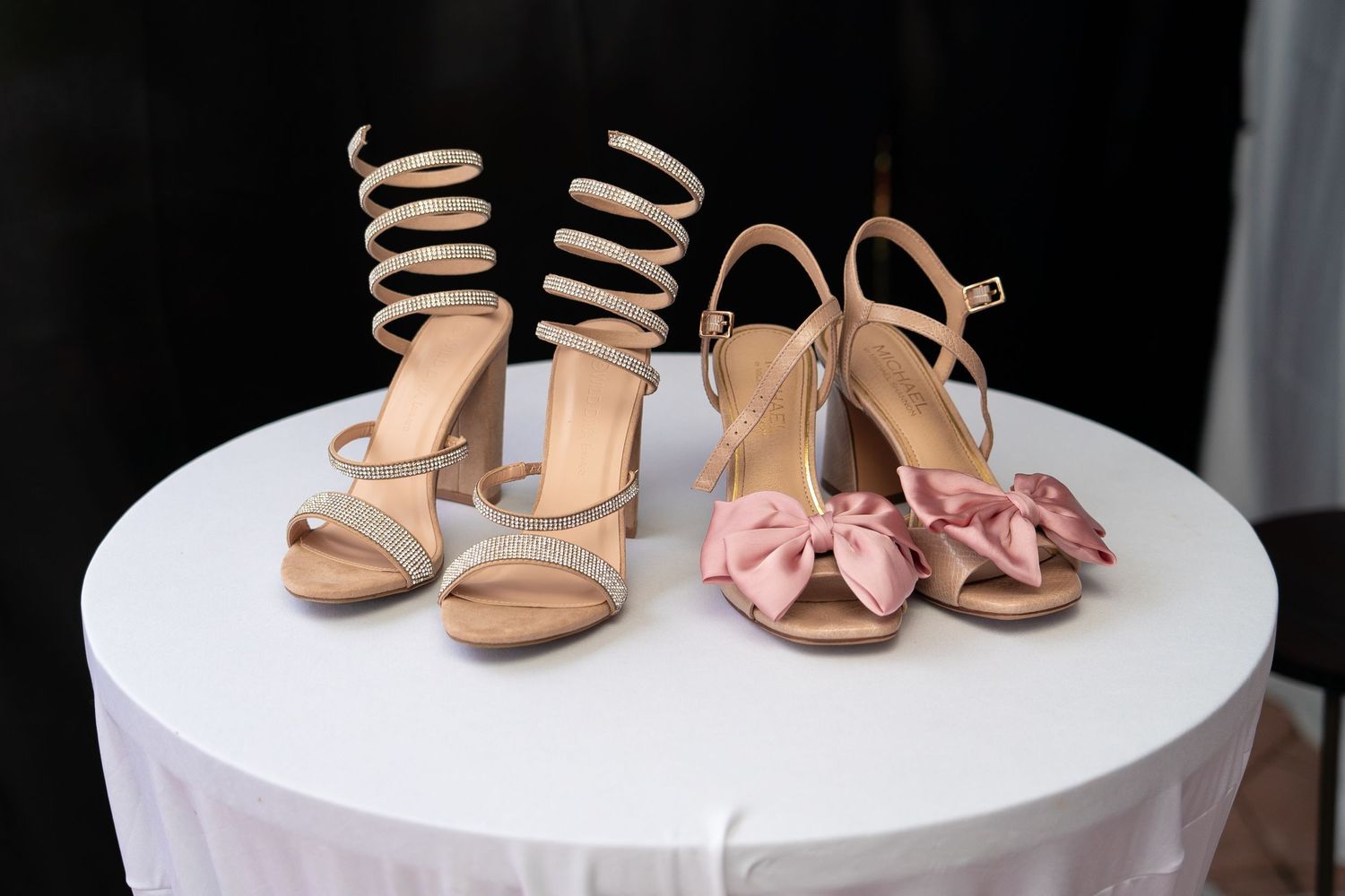 Two pairs of elegant heels displayed on a white table, one gold spiral and one pink bow style.