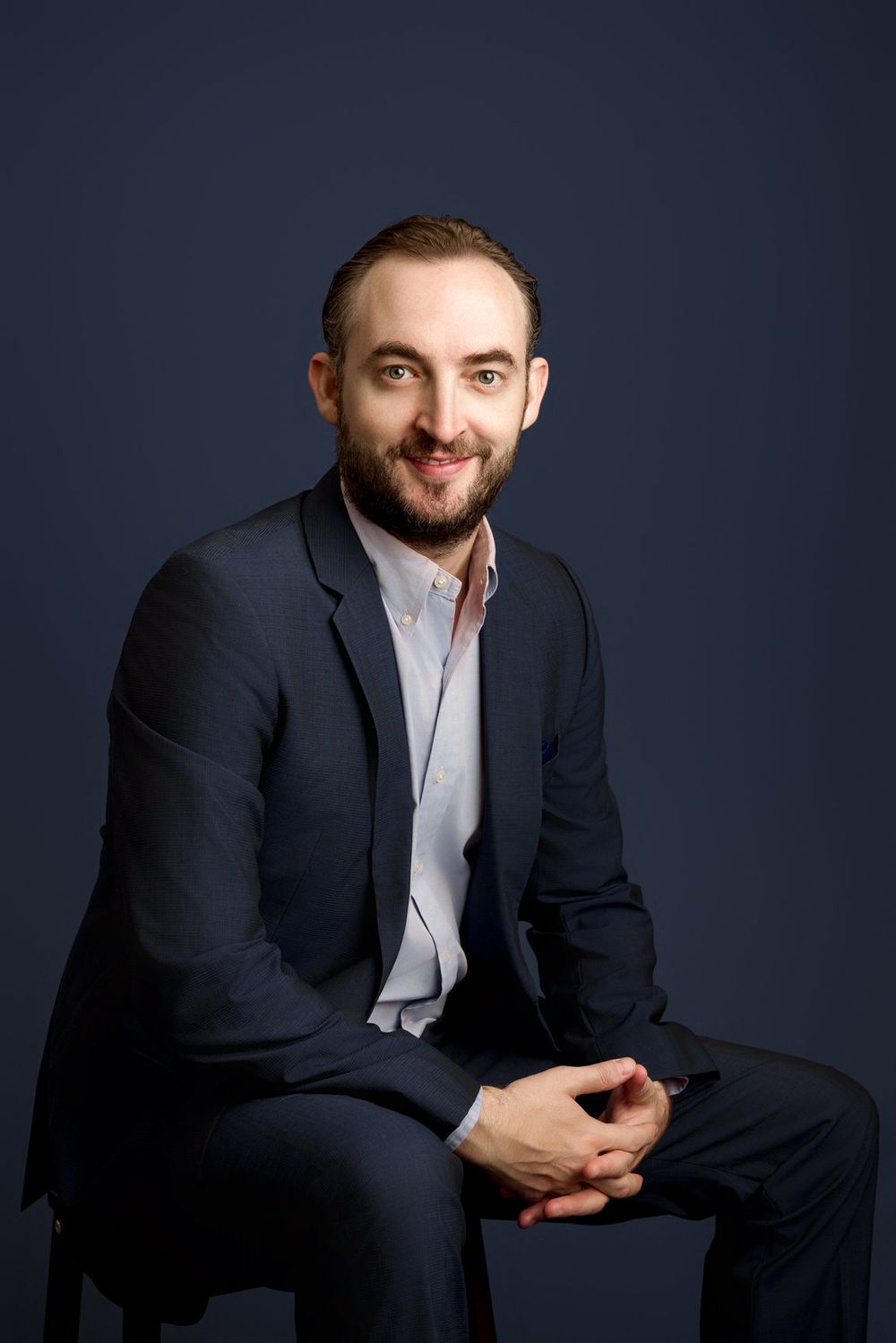 Professional Miami headshot of a seated man in a navy suit against a blue studio background.