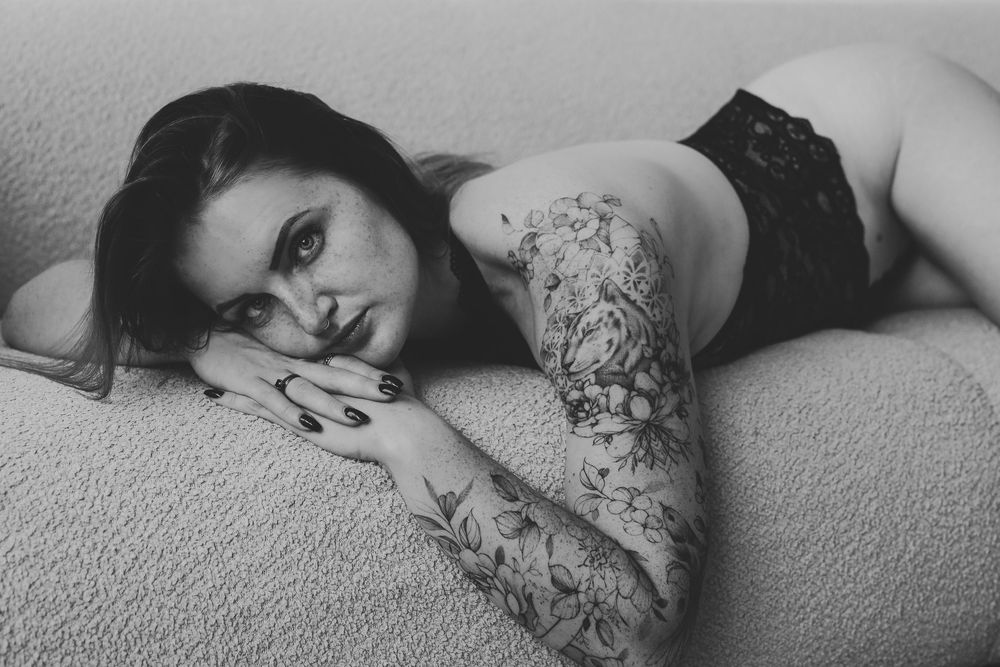 Black and white photo of tattooed woman in lingerie lying on a couch in a boudoir style portrait.