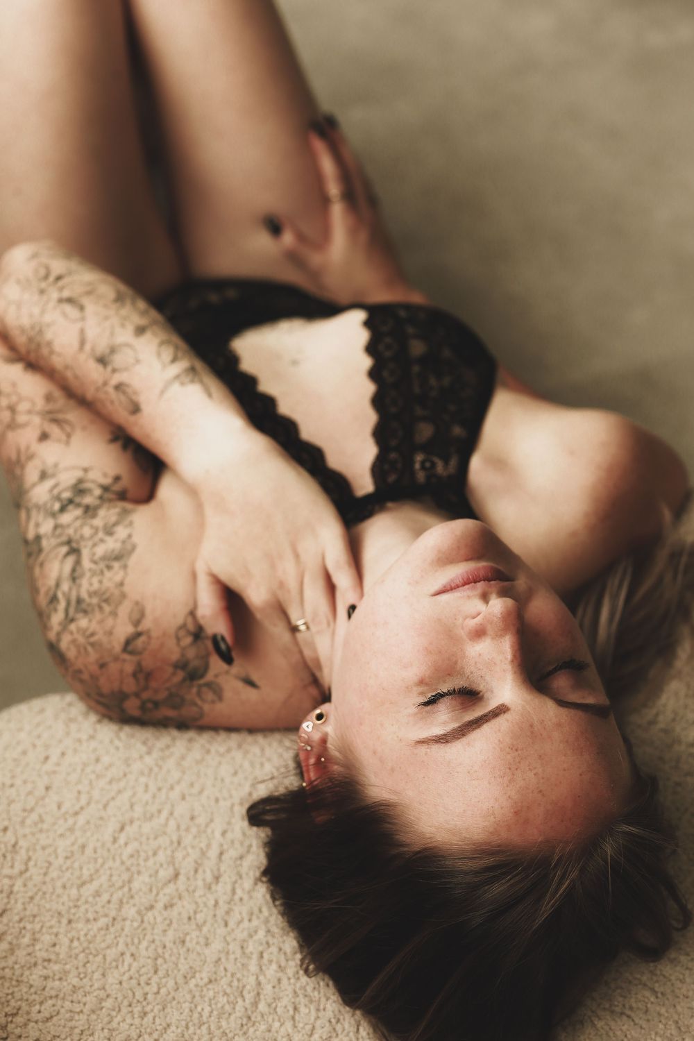 Woman in black lace lingerie lying down, showcasing tattoos on arms and legs in a boudoir photography style.
