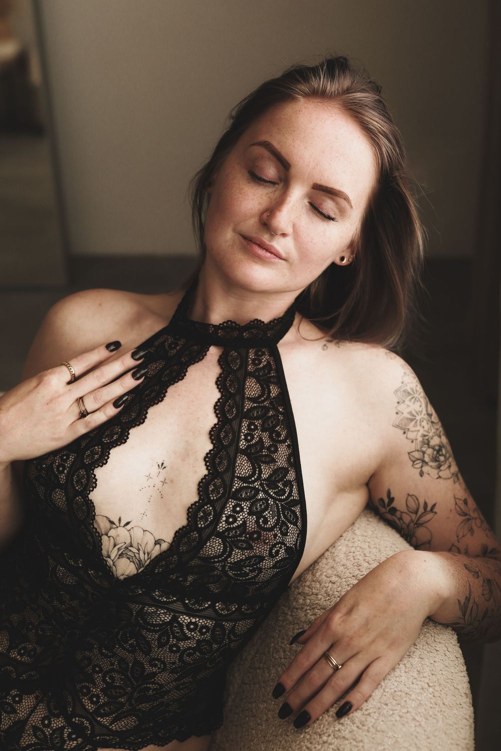 Woman in black lace halter bodysuit with floral tattoos poses gracefully with eyes closed in soft lighting.