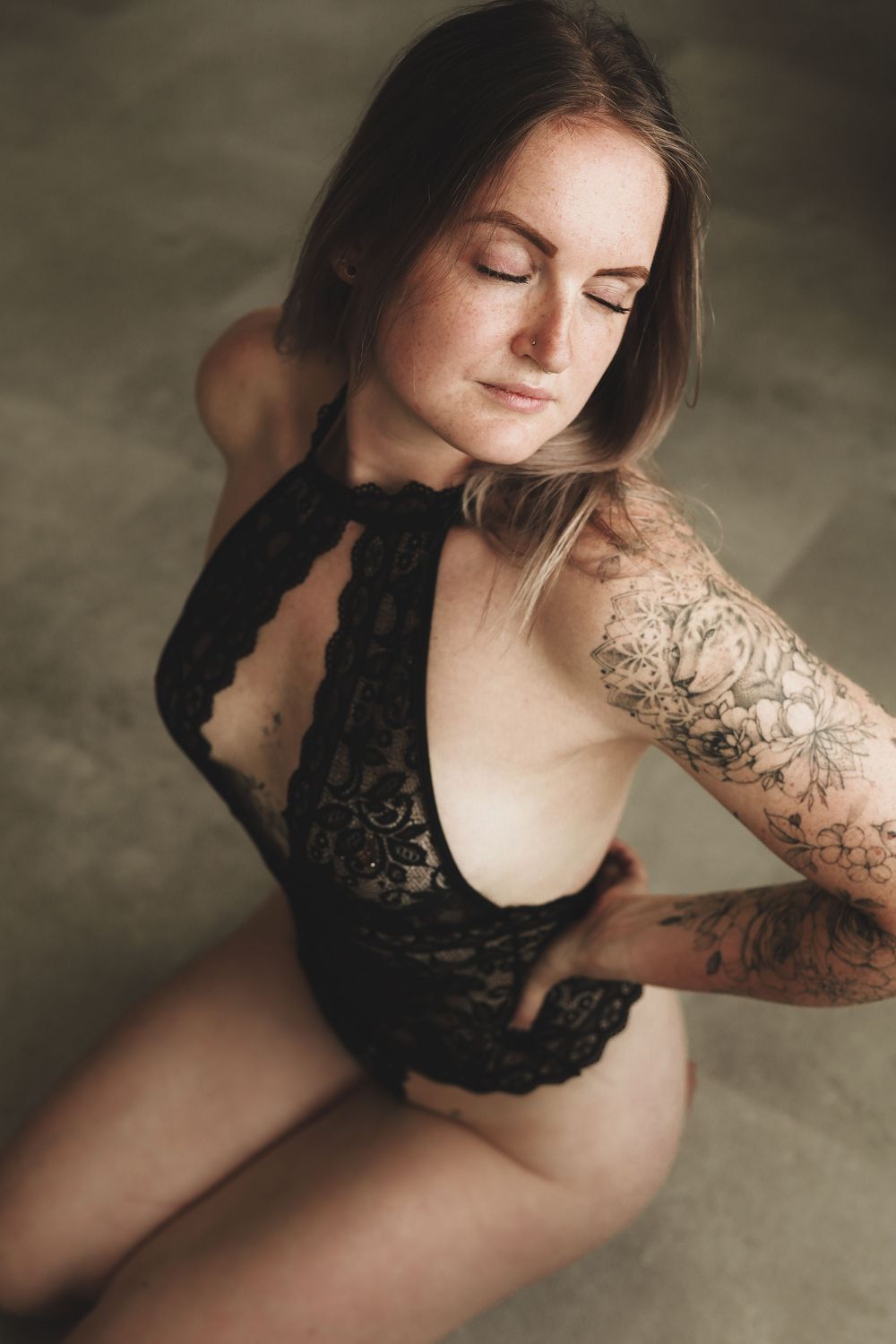 Woman in black lace bodysuit with floral arm tattoo posing gracefully with eyes closed against neutral background.
