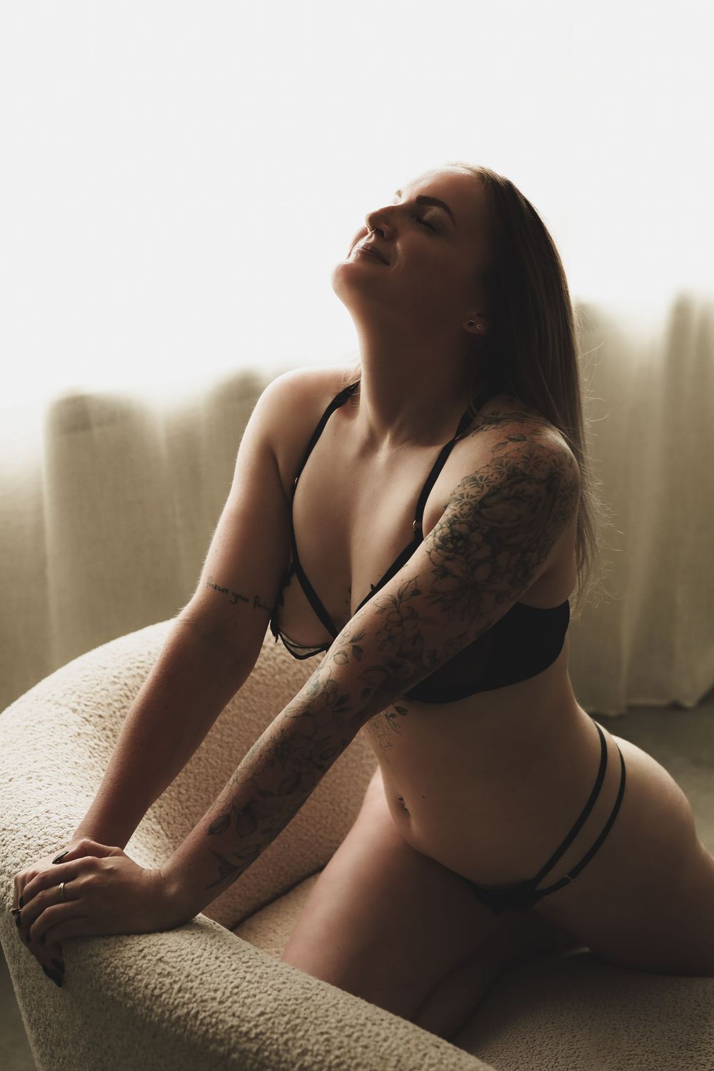 Woman in black lingerie with tattoo sleeve posing gracefully on a couch in soft natural light.