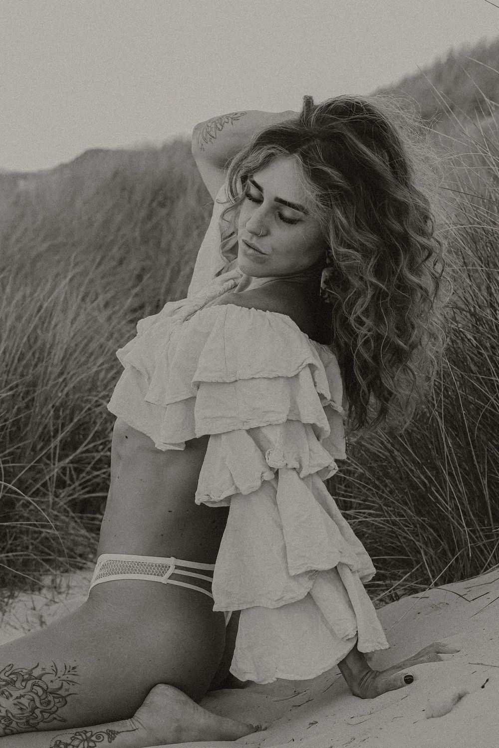 Black and white photo of a woman in ruffled off-shoulder top sitting on sandy beach dunes with tall grass behind her.