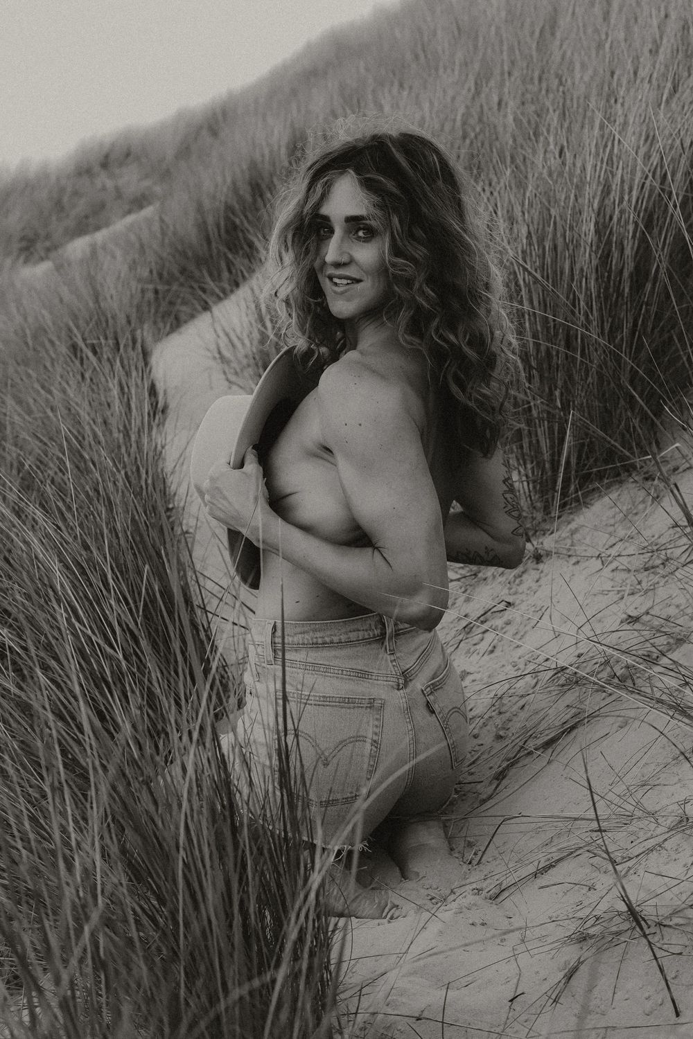 Black and white photo of curly-haired woman posing topless in sandy beach dunes with tall grass.