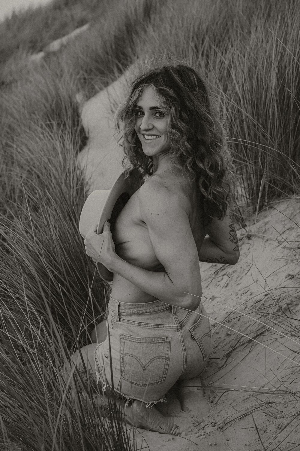 Black and white photo of a smiling woman with curly hair posing on sandy beach dunes wearing denim shorts.