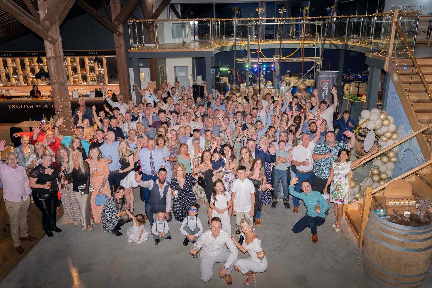 A large group of wedding guests dancing and celebrating together on a rustic wooden dance floor at a reception venue.